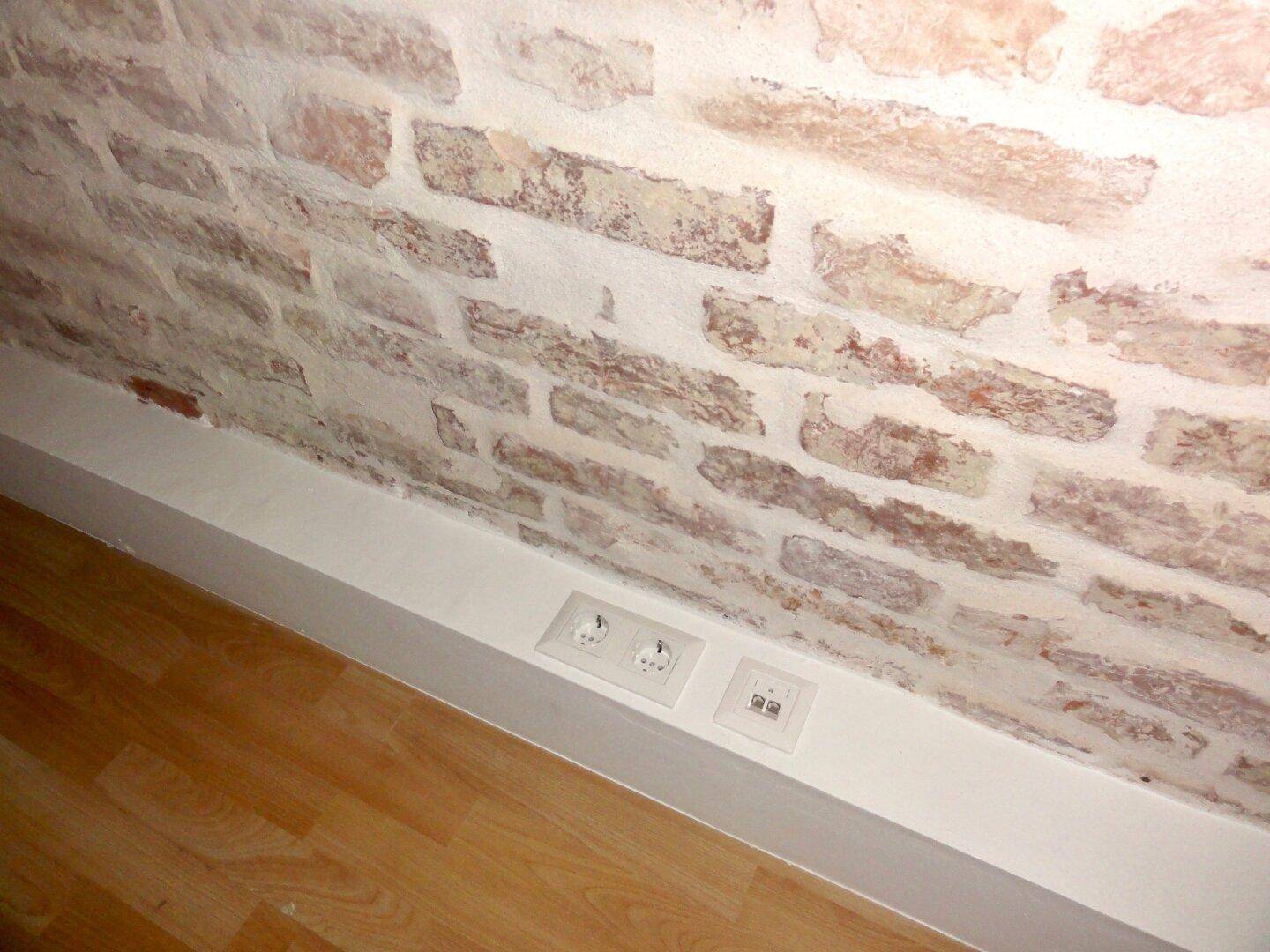 Detailaufnahme einer freigelegten Ziegelwand mit Steckdosen über dem Holzboden.