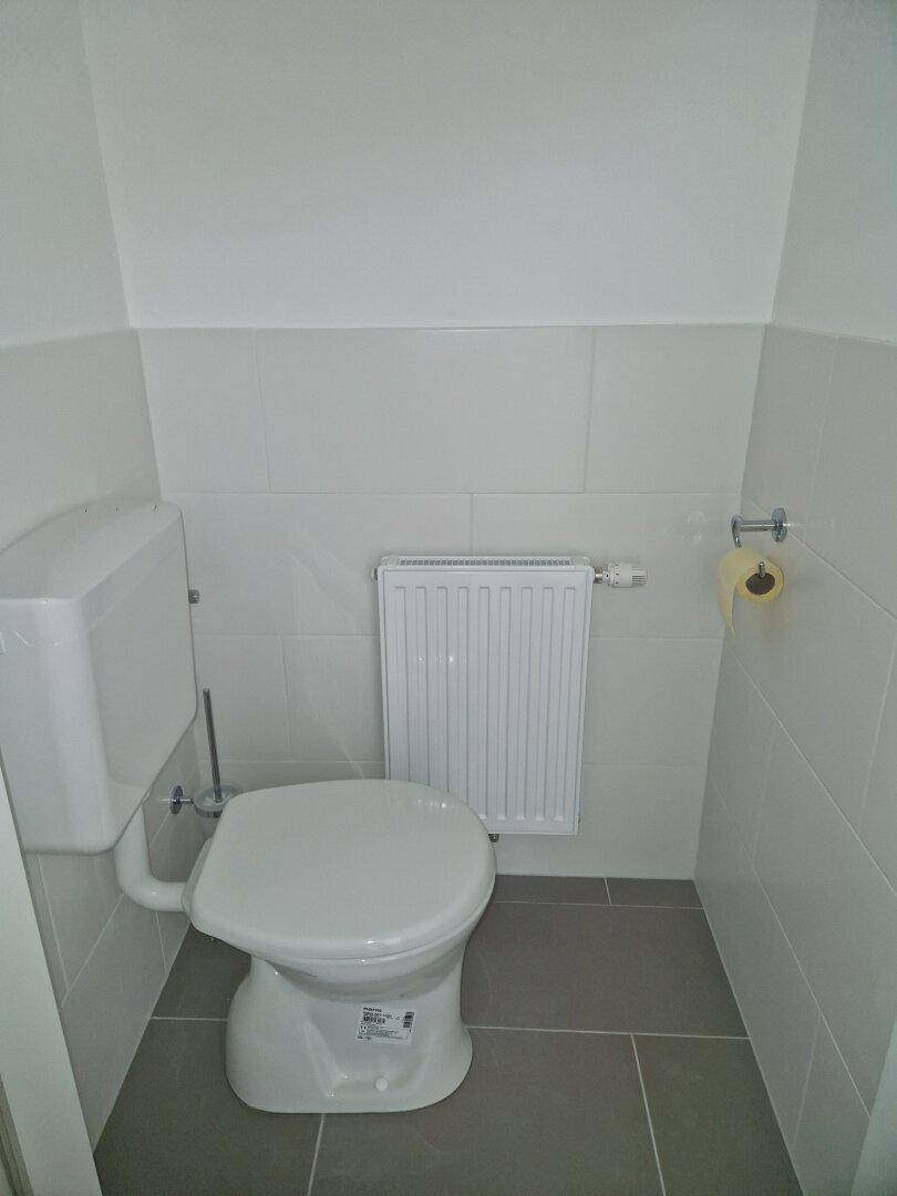 Separates WC mit weißer Toilette, Heizkörper und hellen Fliesen an den Wänden.