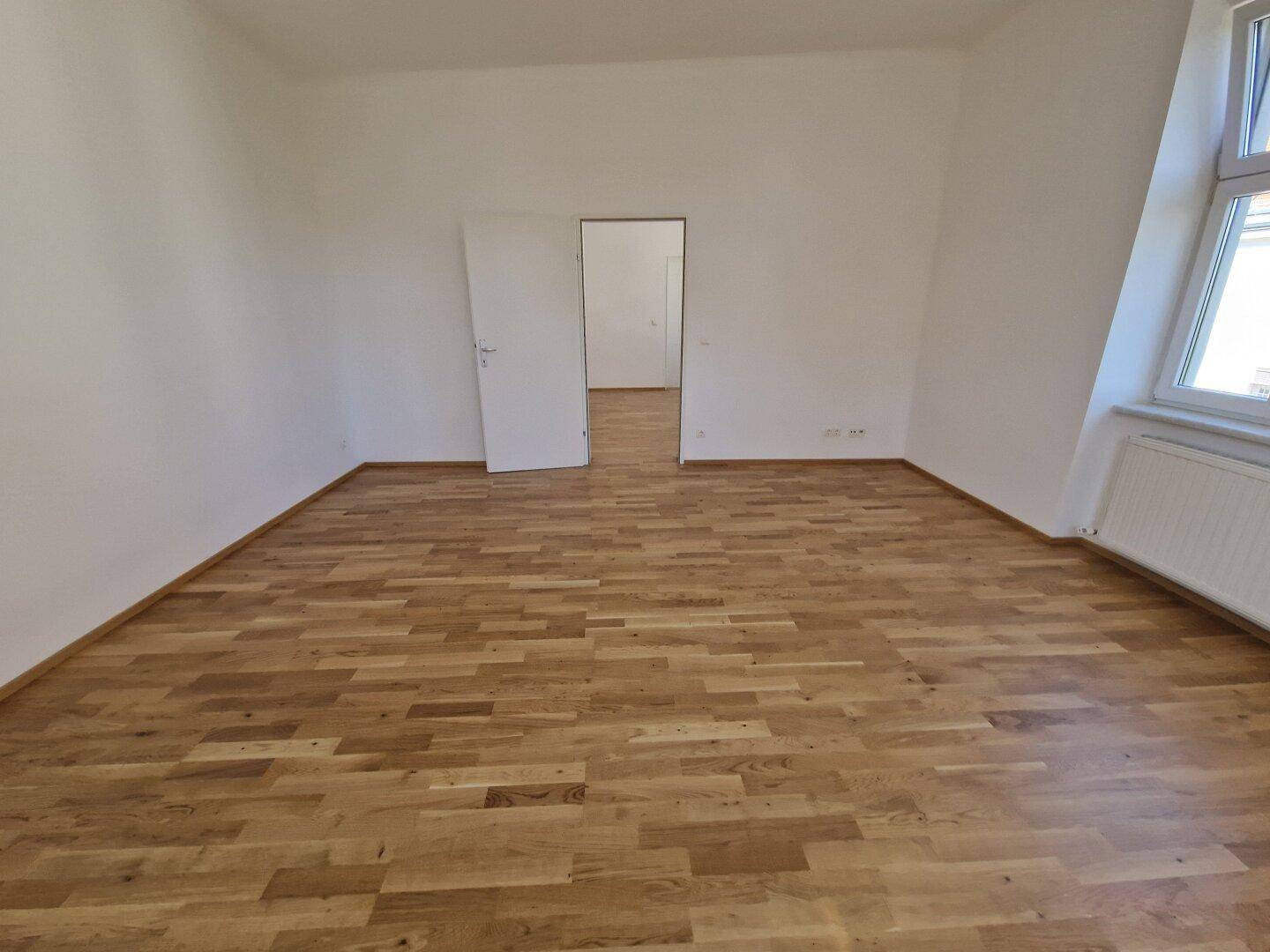 Geräumiges, unmöbliertes Zimmer mit hellem Holzparkett und viel Tageslicht durch das Fenster.
