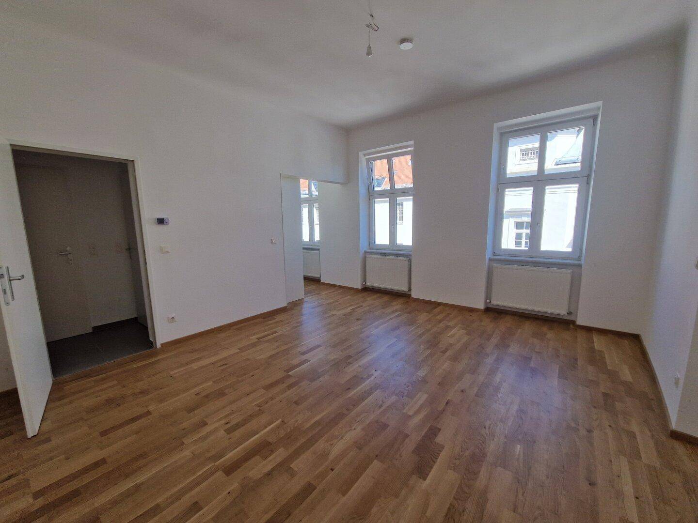 Lichtdurchfluteter Raum mit zwei großen Fenstern, neuem Parkettboden und Zugang zu weiteren Zimmern.