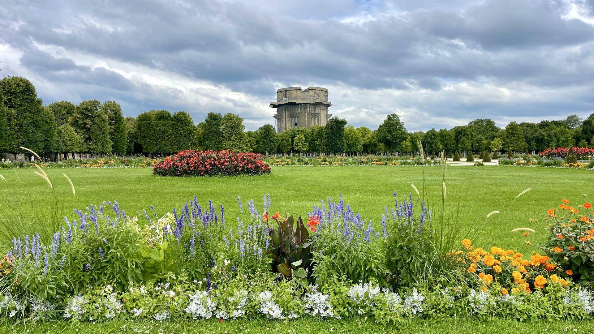 Weitläufiger Park mit gepflegten Rasenflächen, bunten Blumenbeeten und einem historischen Flakturm im Hintergrund.