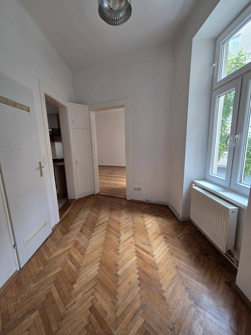 Helles Zimmer mit Fischgrätparkett und hohen Fenstern, bietet Zugang zu weiteren Räumen.