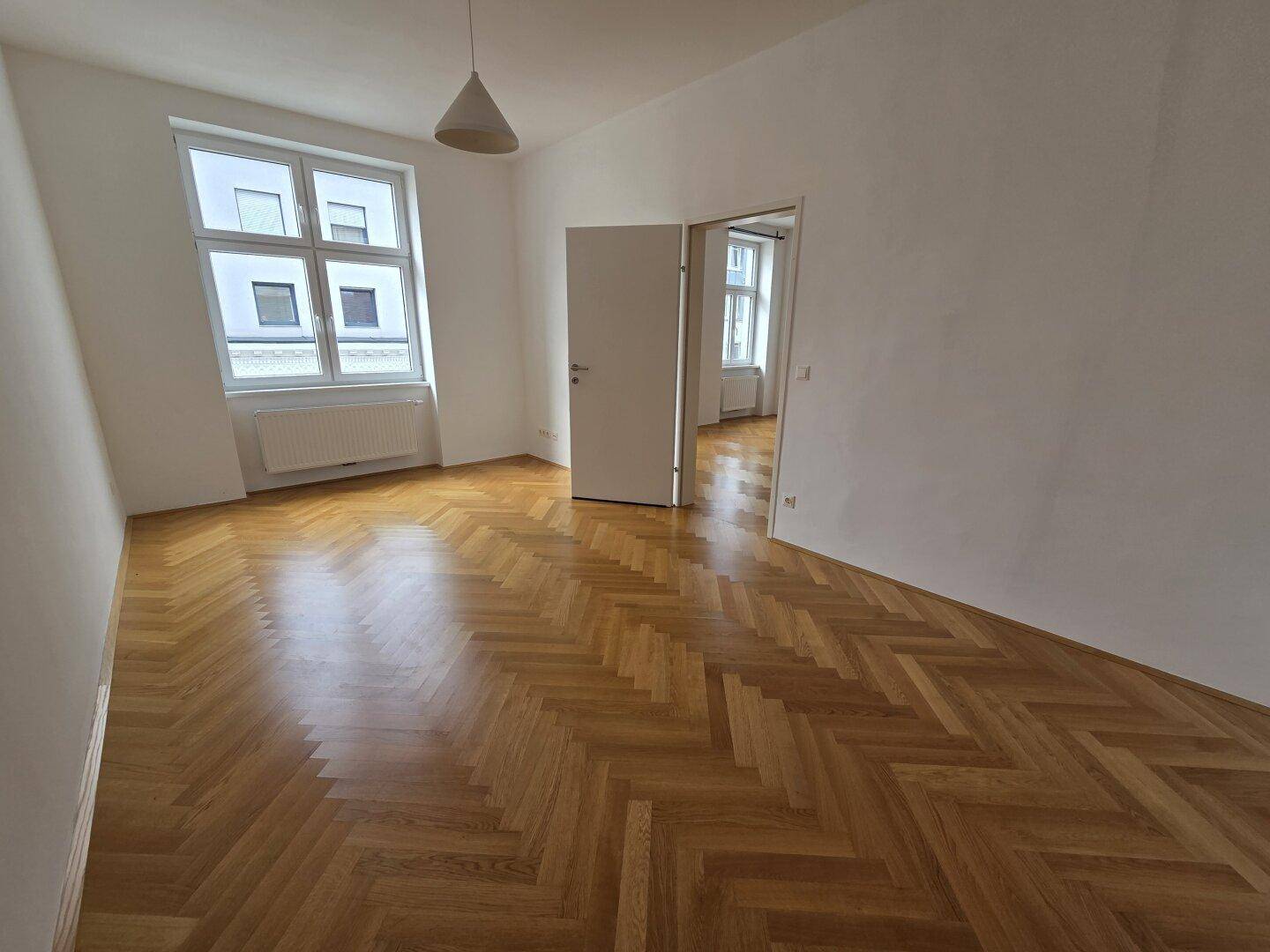 Geräumiges Zimmer mit klassischem Fischgrätparkett und viel Tageslicht durch große Fenster.
