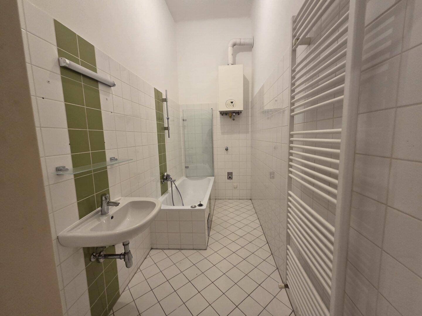 Badezimmer mit Badewanne, Waschbecken und grün-weißen Fliesen, ausgestattet mit Handtuchheizkörper.