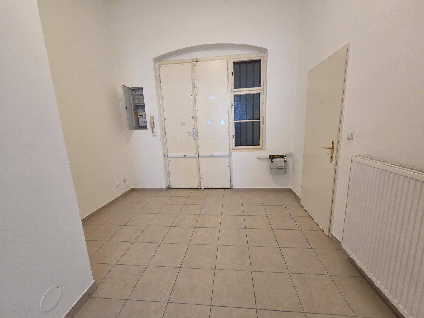 Heller Eingangsbereich mit Fliesenboden, zwei Türen und einem Fenster mit Gitter, inklusive Heizkörper.