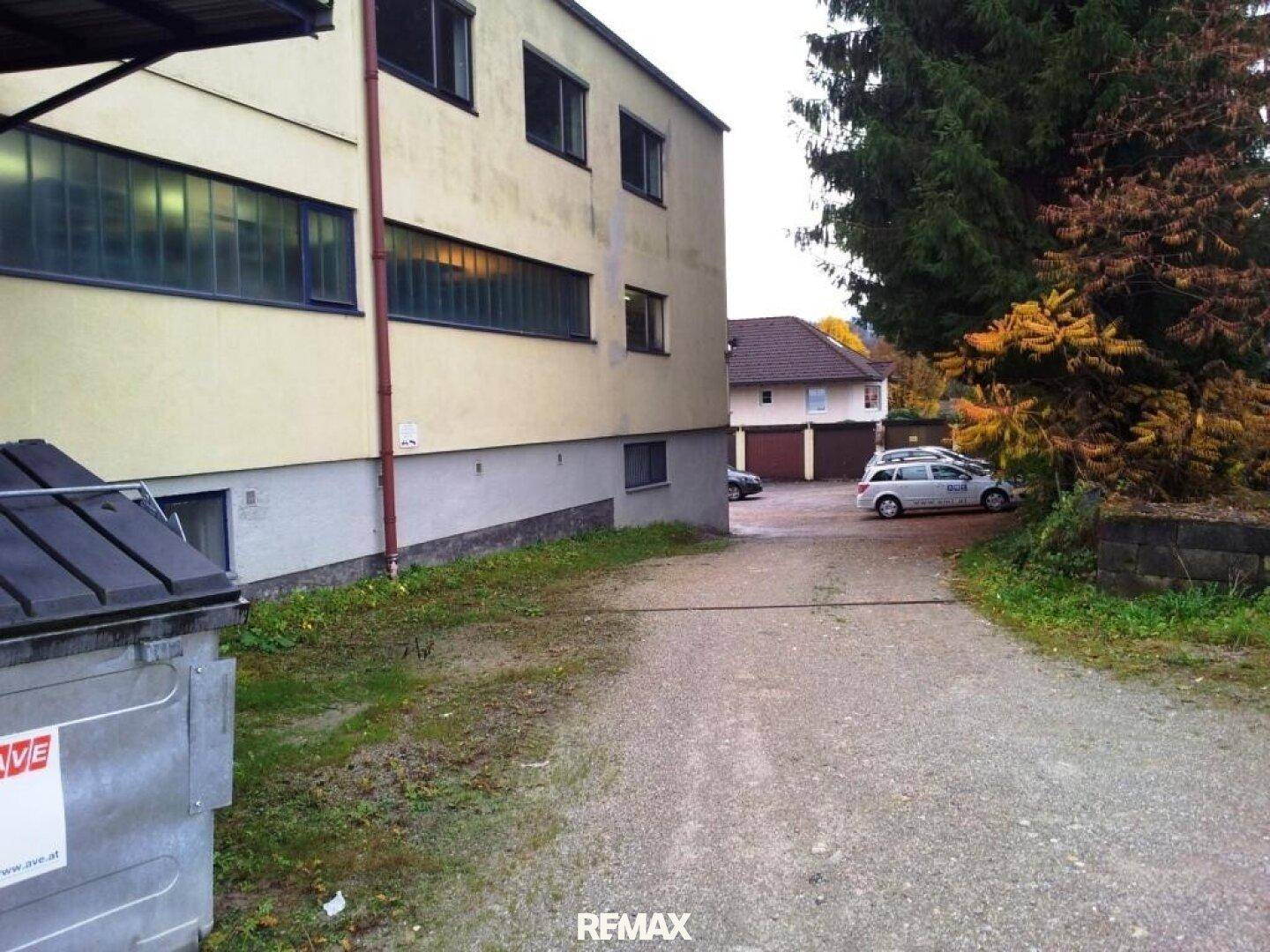 Rückseite des Gebäudes mit Schotterweg, Bäumen und parkenden Autos im Hintergrund.
