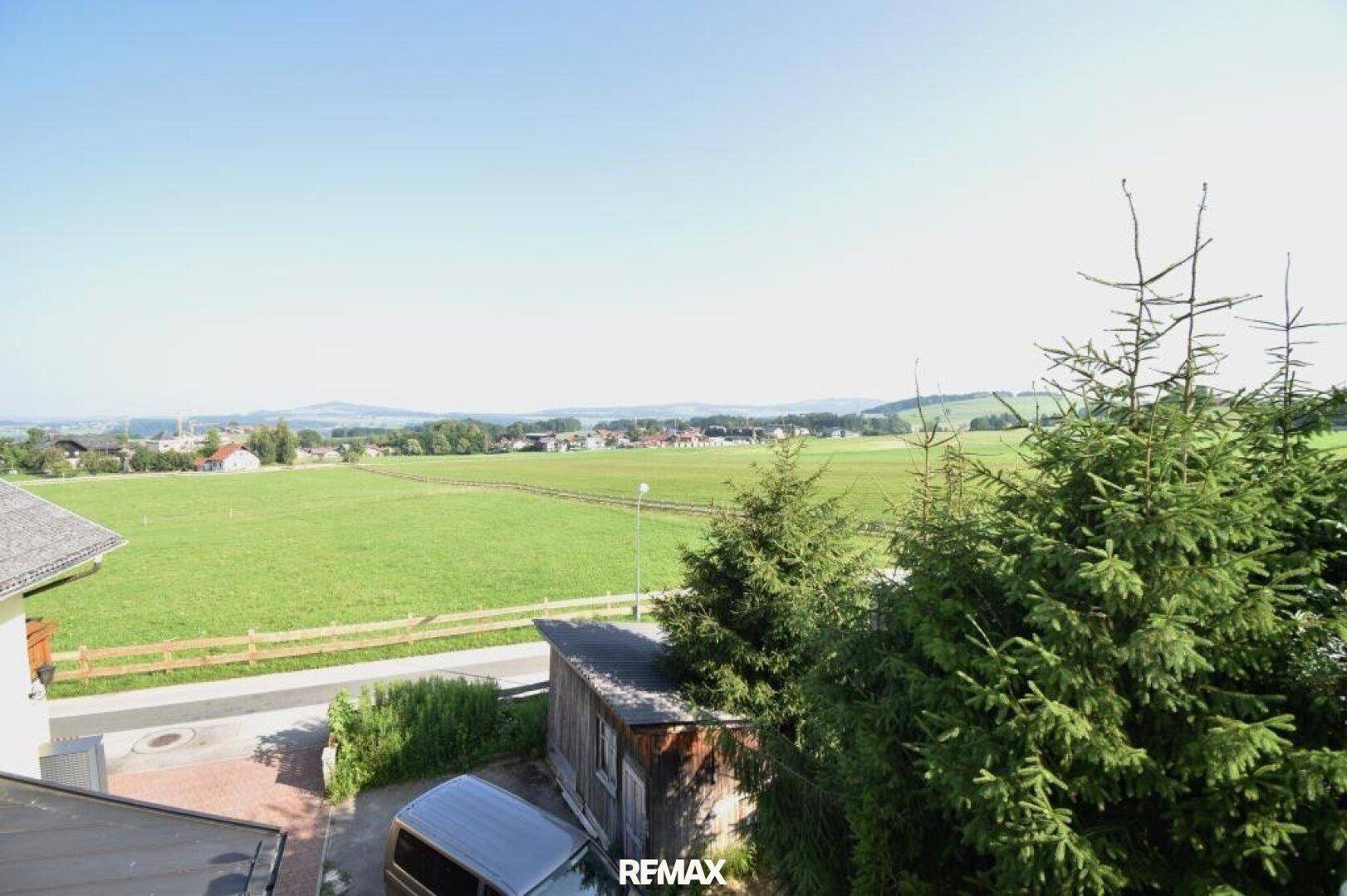 Weitläufiger Blick über grüne Felder, ländliche Umgebung und ferne Berge unter blauem Himmel.