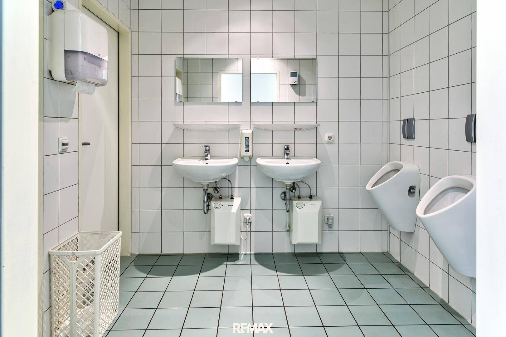Geräumiger Toilettenraum mit zwei Waschbecken und zwei Urinalen, komplett gefliest in Weiß und Hellblau.