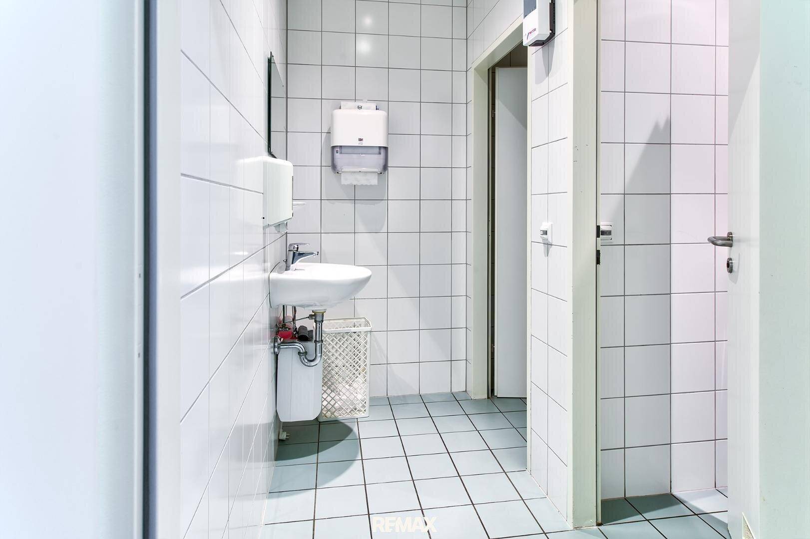 Gefliester Toilettenraum mit Waschbecken, Spiegel und Papierhandtuchspender, in hellem Design.