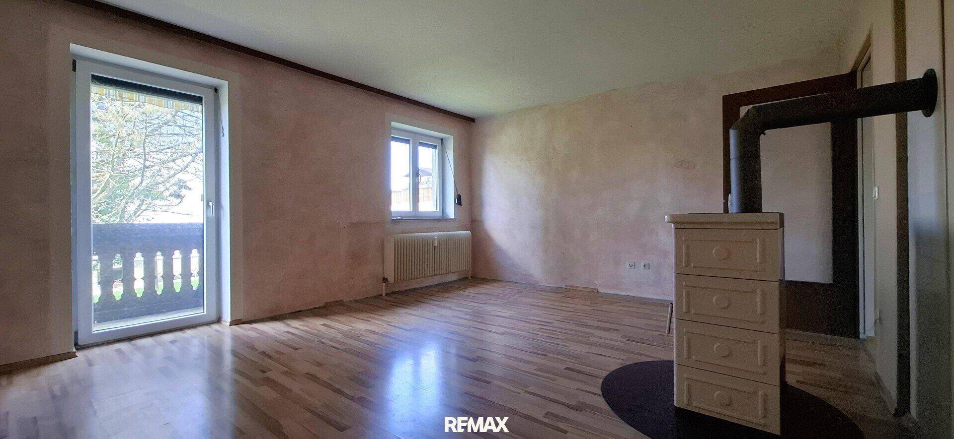 Helles Wohnzimmer mit Holzboden, Kachelofen und Zugang zu einem Balkon durch eine Fenstertür.