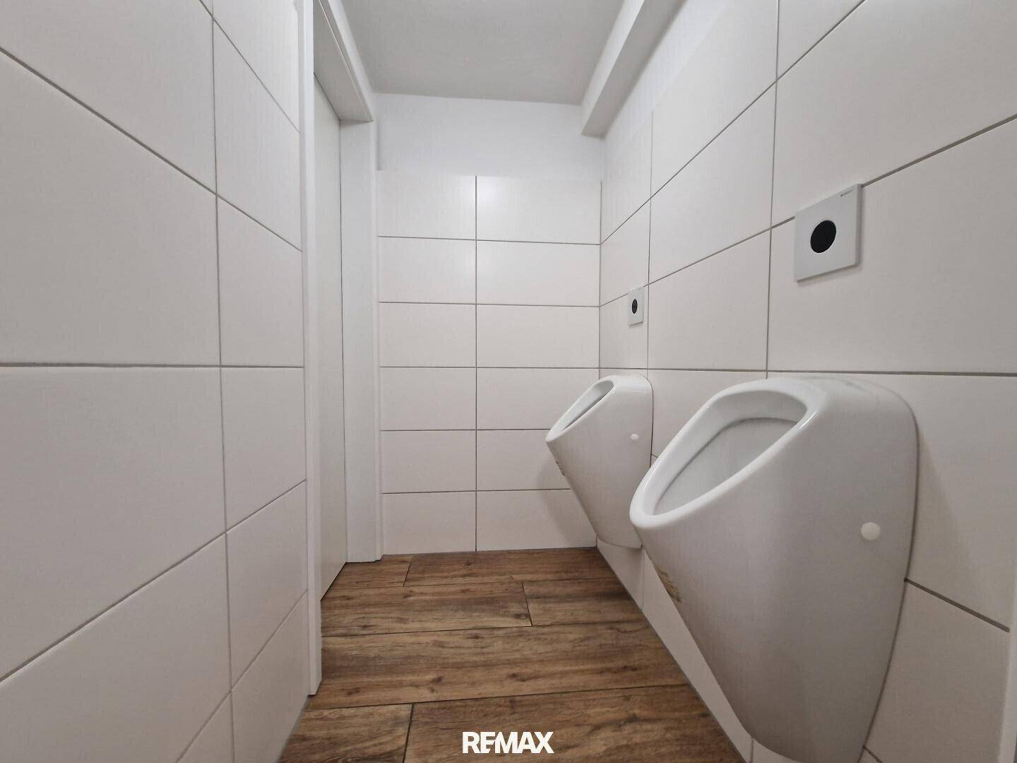 Sauberer Herrenwaschraum mit zwei Urinalen, weißen Fliesenwänden und Holzoptik-Boden.