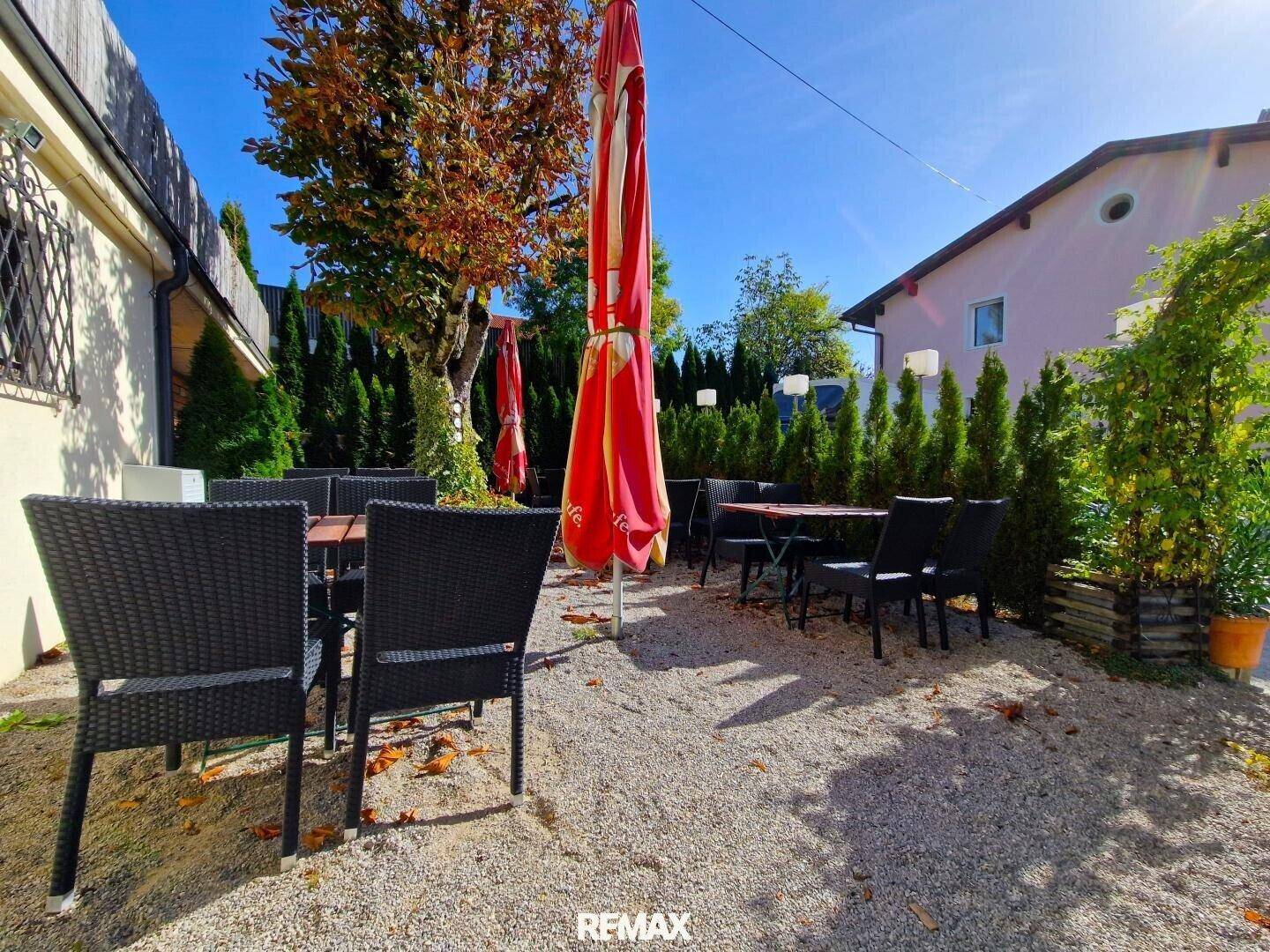 Sonniger Außenbereich mit Tischen und Stühlen unter großen roten Sonnenschirmen, umgeben von viel Grün.