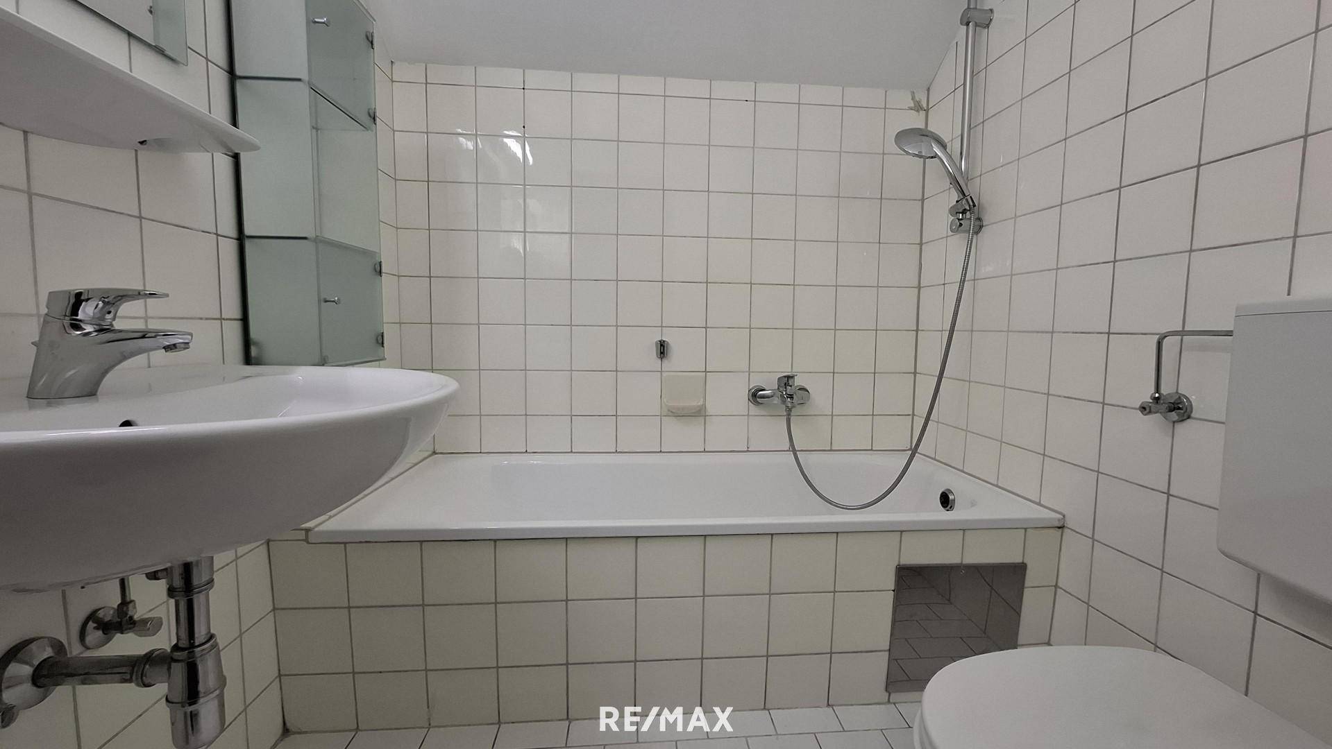Funktionales Badezimmer mit weißen Fliesen, Badewanne mit Duschfunktion, Waschbecken und Toilette.