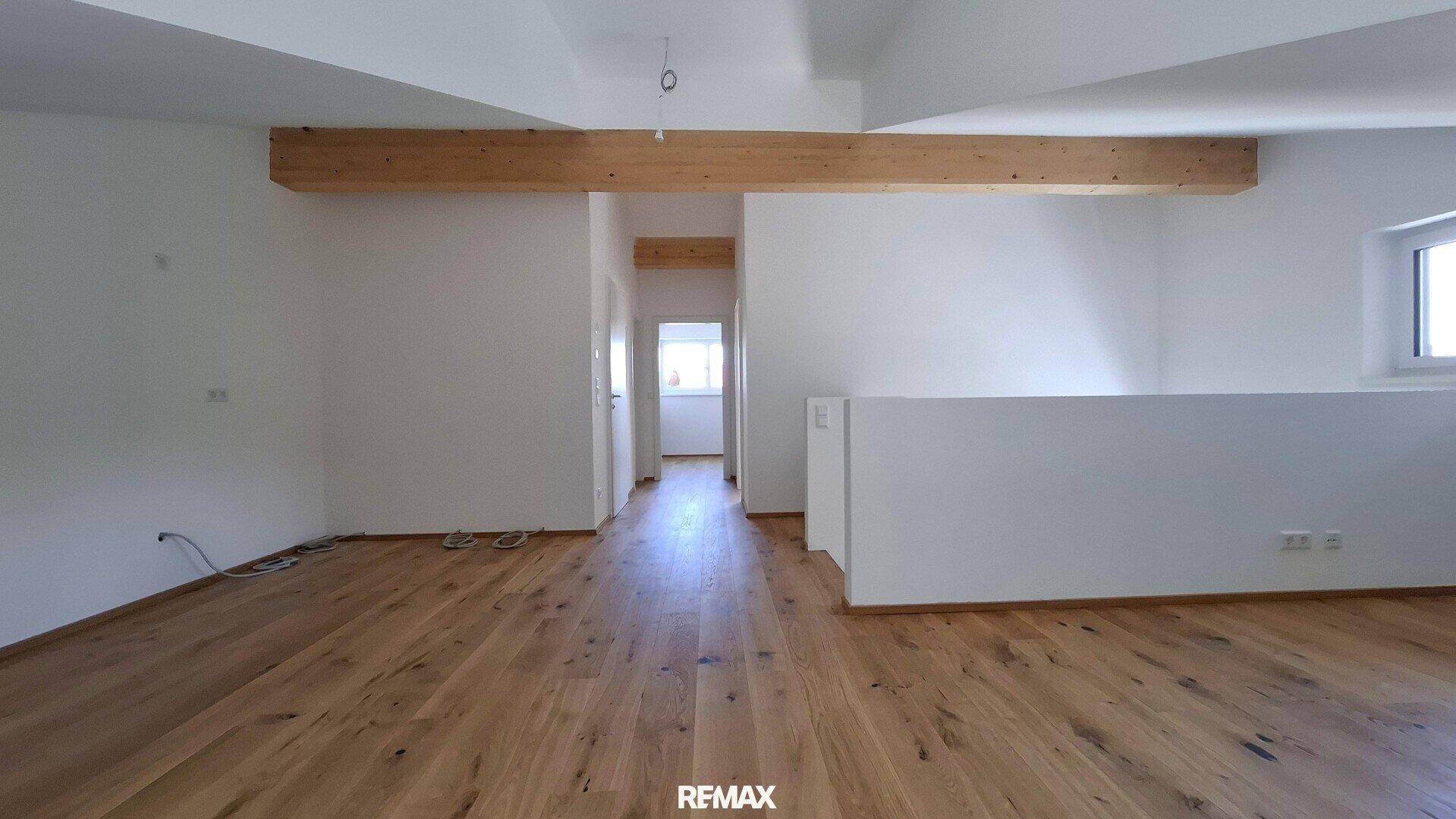 Offener Wohnbereich mit Holzboden und sichtbarem Holzbalken, der zu weiteren Räumen führt.