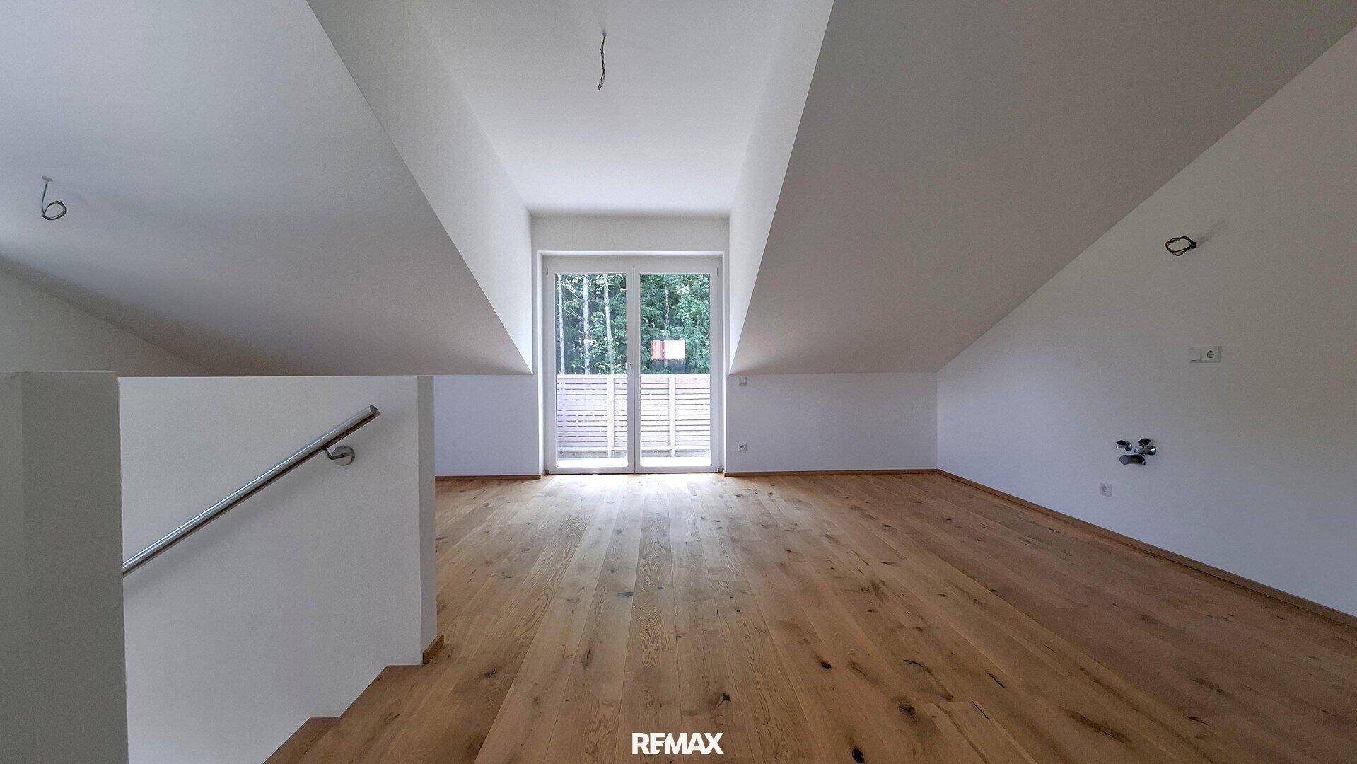 Geräumiger, heller Dachgeschossraum mit Holzboden und großer Glastür, die viel Licht hereinlässt.