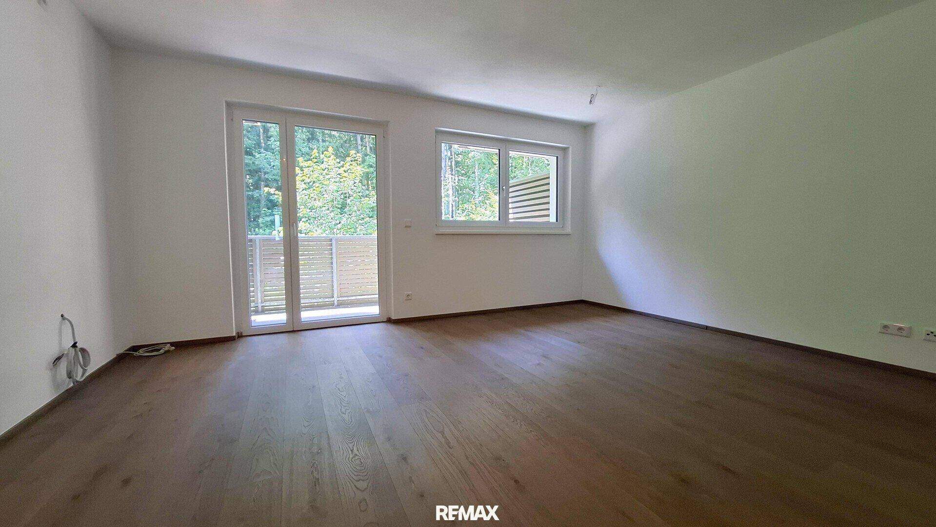 Heller, leerer Raum mit dunklem Holzfußboden, weißen Wänden und Zugang zu einem Balkon oder einer Terrasse.