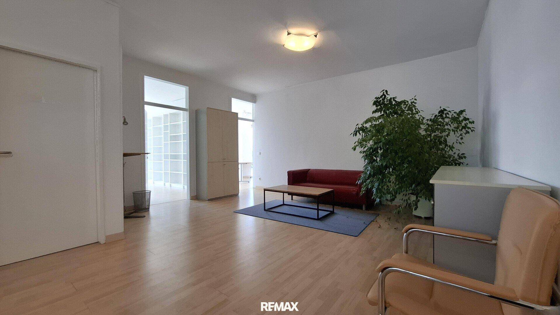 Wartebereich mit rotem Sofa, Couchtisch und einer großen Pflanze, ergänzt durch Holzboden.