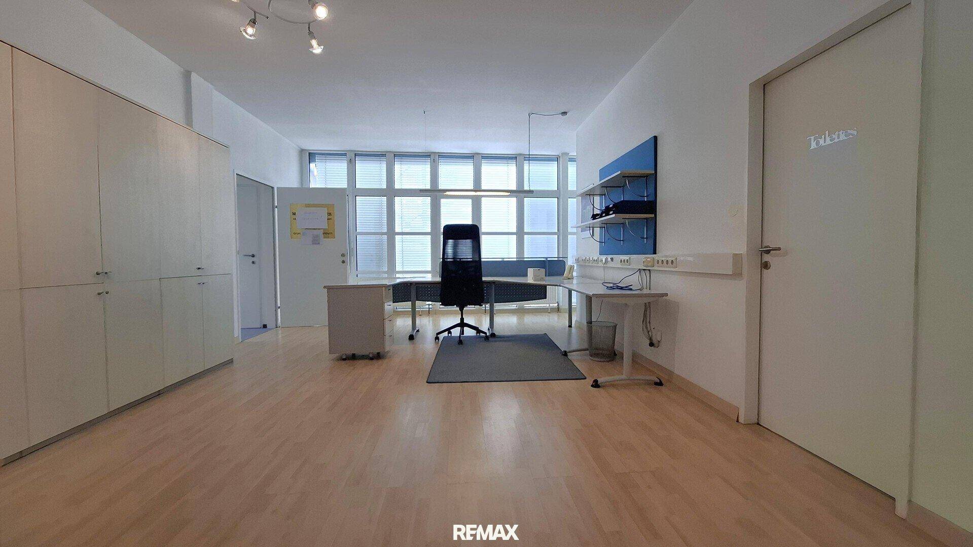 Funktionales Büro mit Schreibtisch, Bürostuhl und Schränken, beleuchtet durch große Fenster.