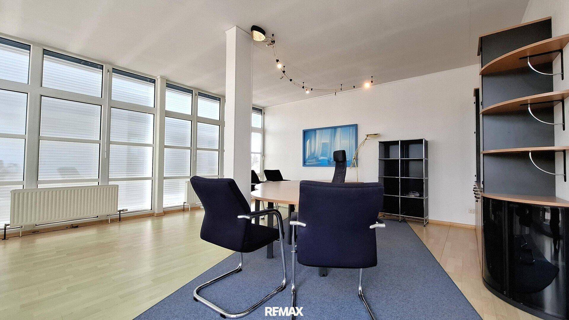 Büro oder Konferenzraum mit großen Fenstern, Holzboden und einem runden Tisch mit Stühlen.