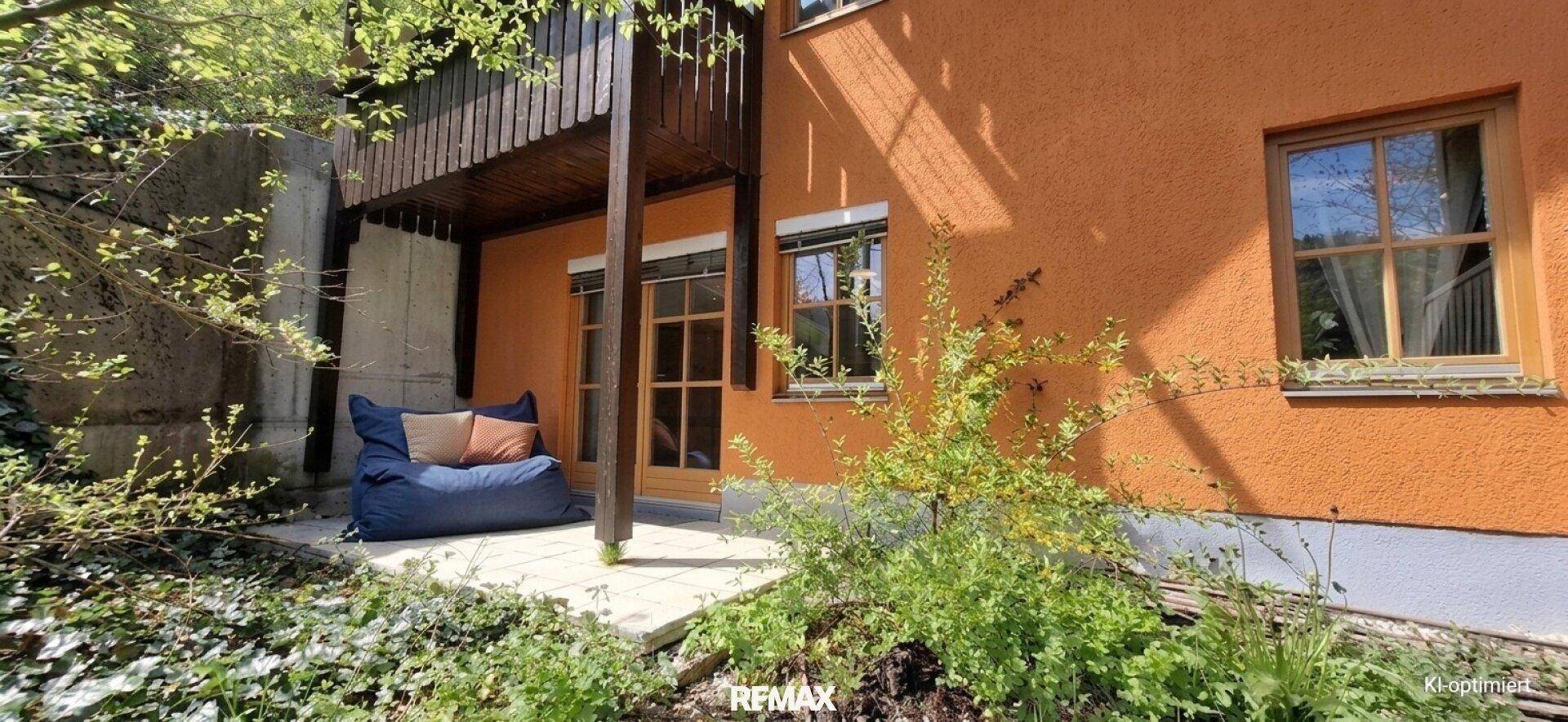 Außenbereich mit kleiner gepflasterter Terrasse, orangefarbener Fassade und üppigem Grün, ideal zum Entspannen.