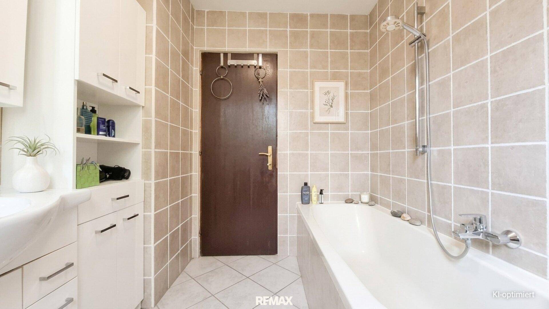 Badezimmer mit beigefarbenen Fliesen, Badewanne und einer Tür, die zu einem weiteren Raum führt.