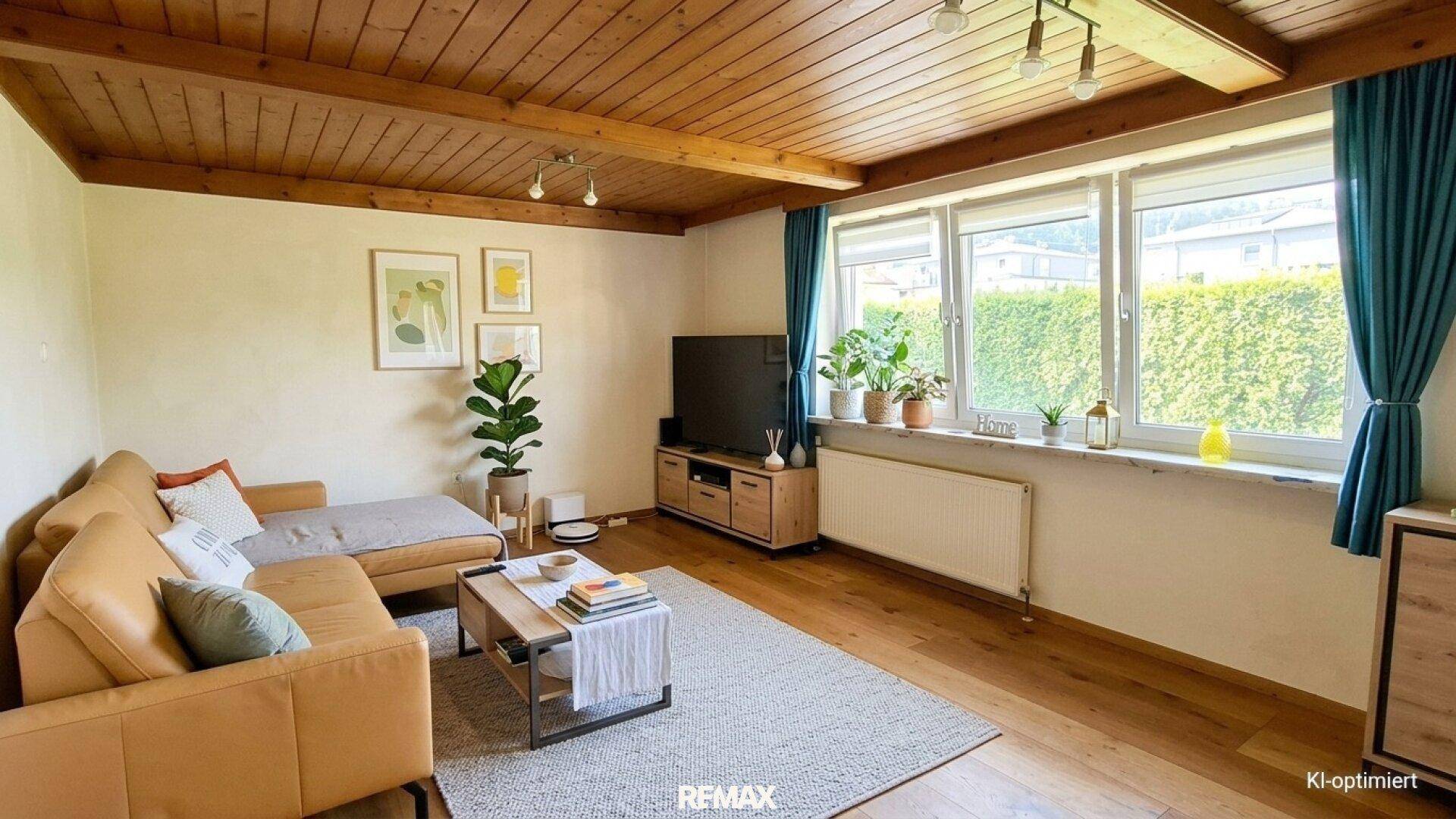 Gemütliches Wohnzimmer mit Holzbalkendecke, bequemer Couch und großen Fenstern mit Heckenblick.