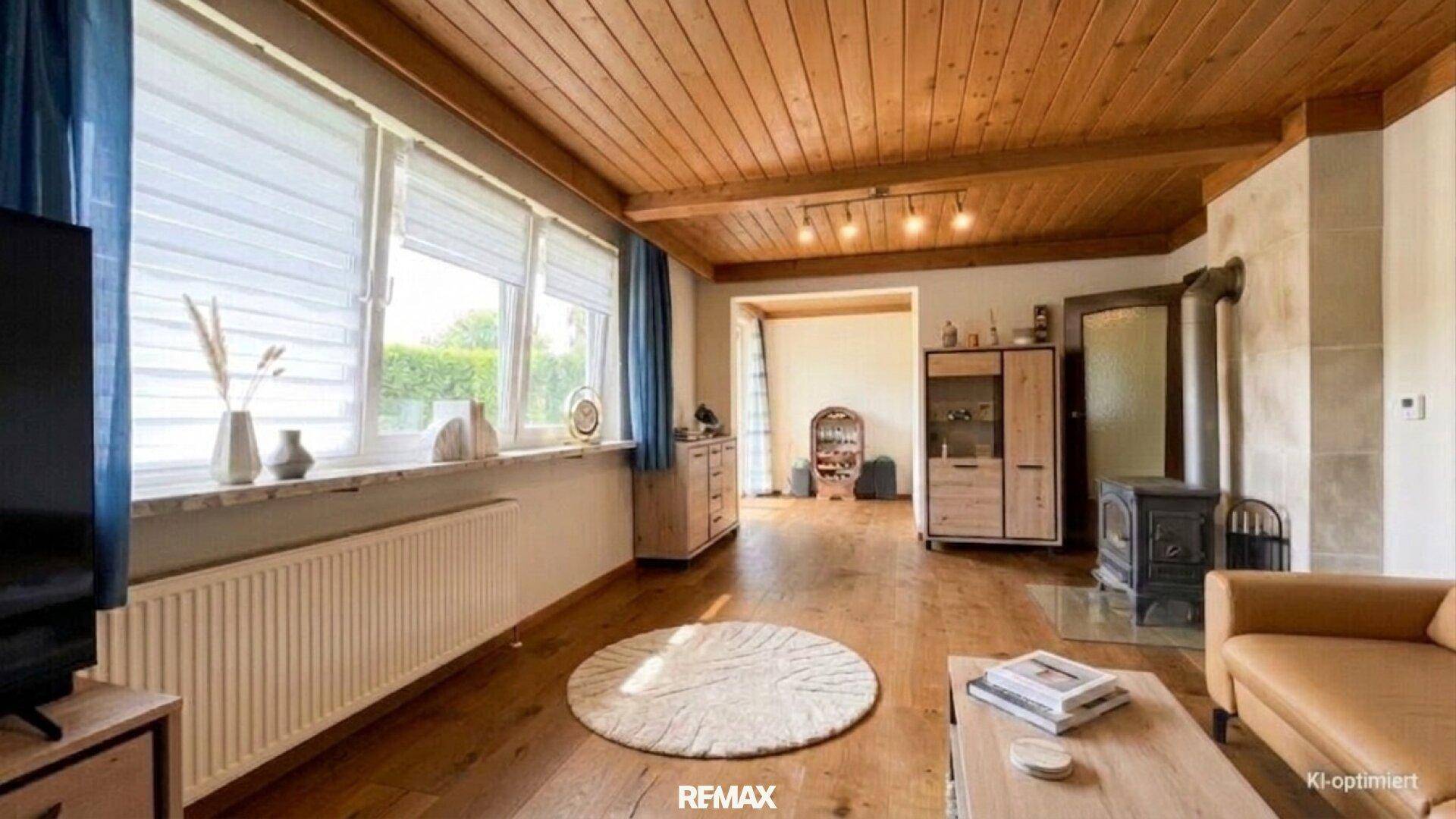 Helles Wohnzimmer mit Holzbalkendecke, Kaminofen und großen Fenstern mit Blick ins Grüne.