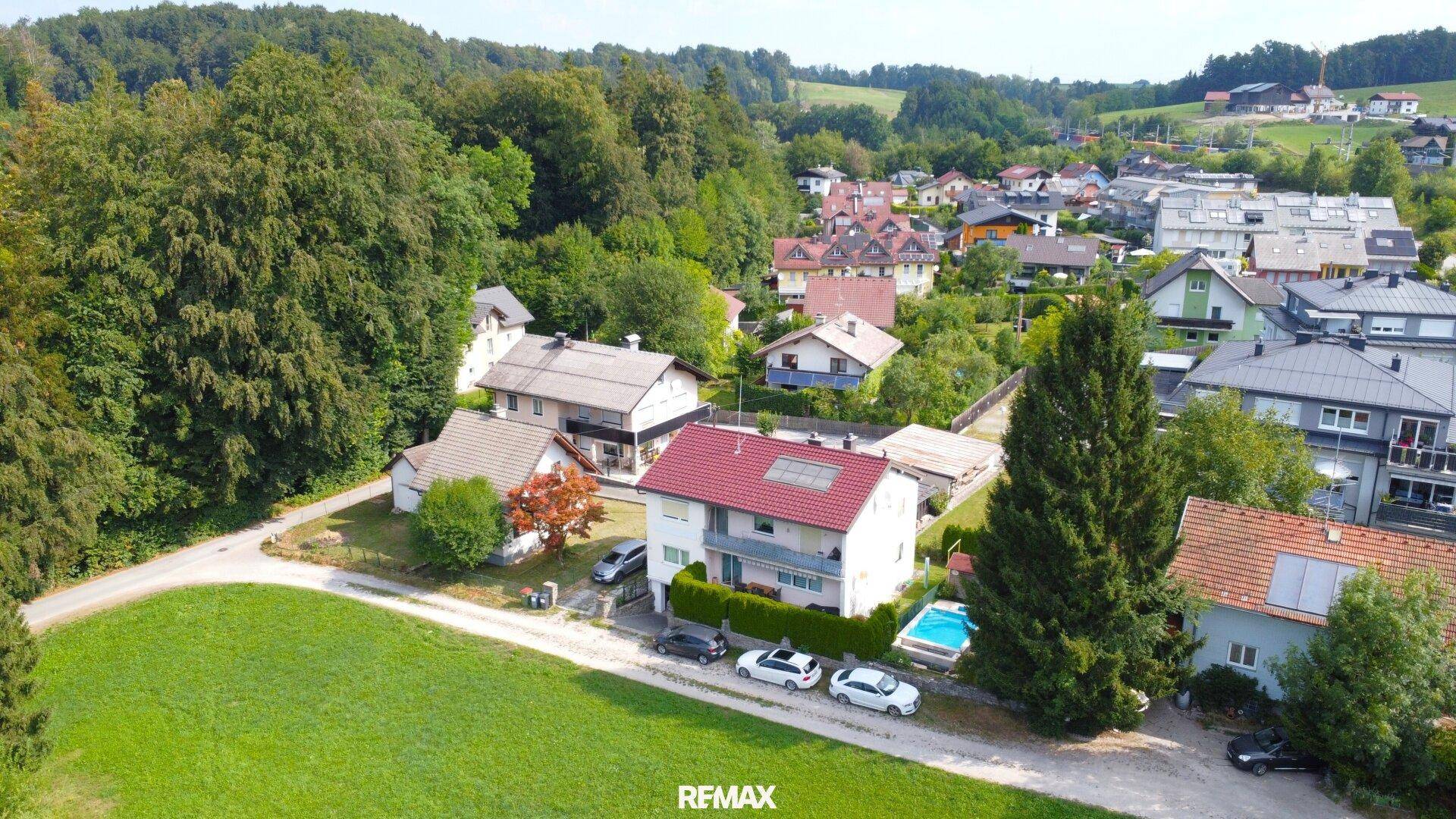 Luftaufnahme eines Einfamilienhauses mit Pool und umliegender Wohnsiedlung in grüner Umgebung.
