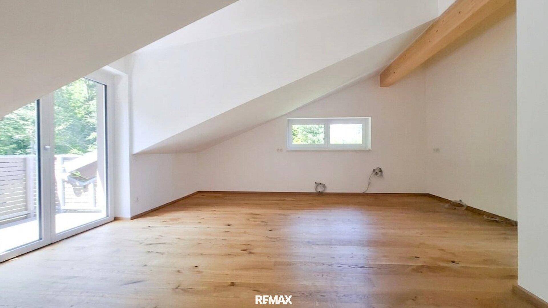 Heller, leerer Raum mit Holzboden, Dachschrägen und großem Fenster zum Außenbereich.
