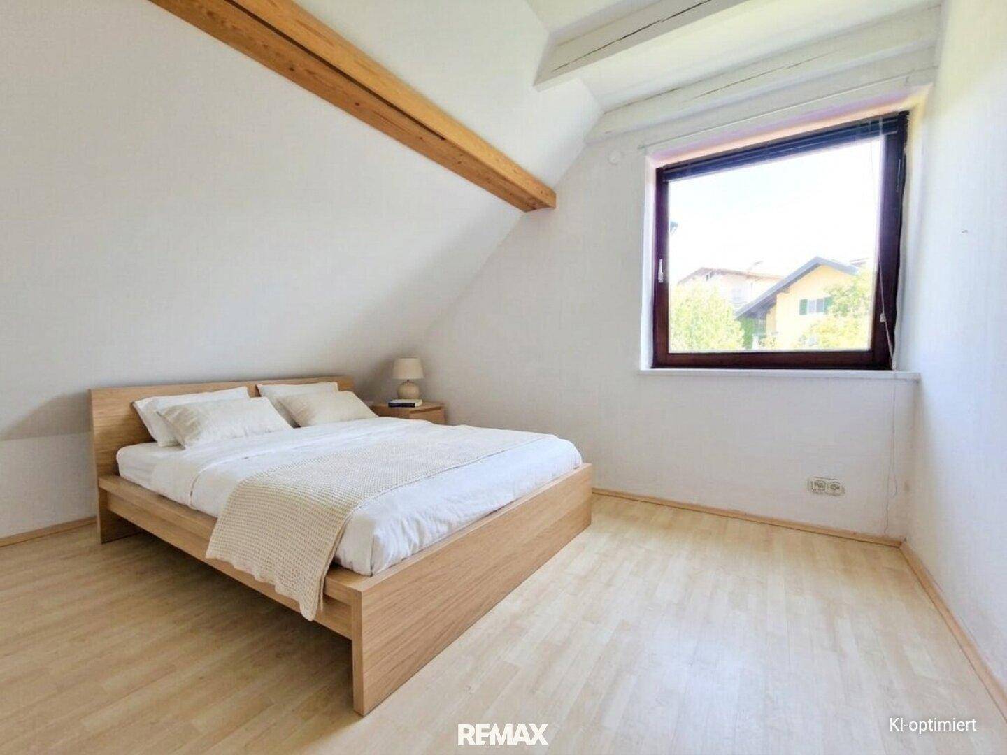 Helles Schlafzimmer mit Holzbett, sichtbarem Holzbalken und einem Fenster mit Blick ins Grüne.