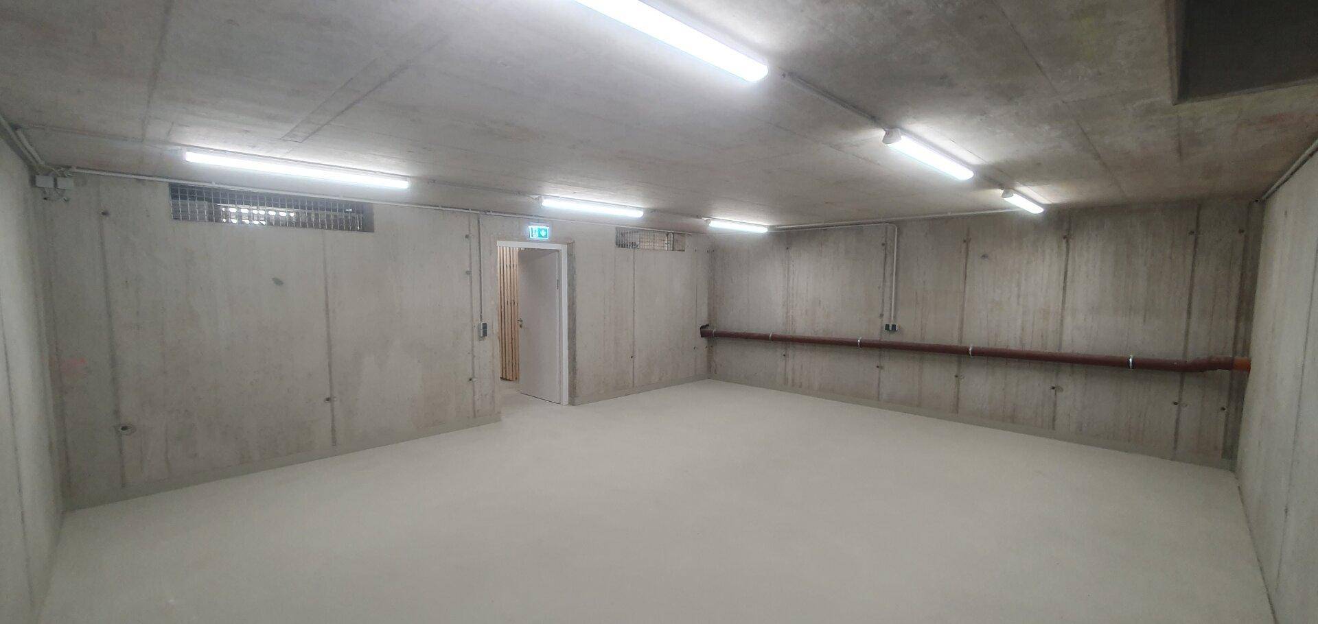 Geräumiger Kellerraum mit unverputzten Betonwänden und hellem Boden, beleuchtet von Neonröhren.
