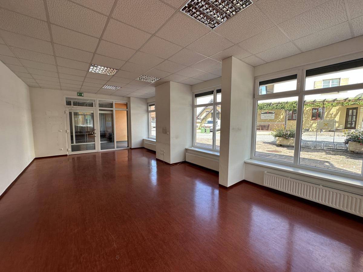 Geräumiger, leerer Gewerberaum mit großen Fenstern, braunem Boden und Rasterdeckenbeleuchtung.