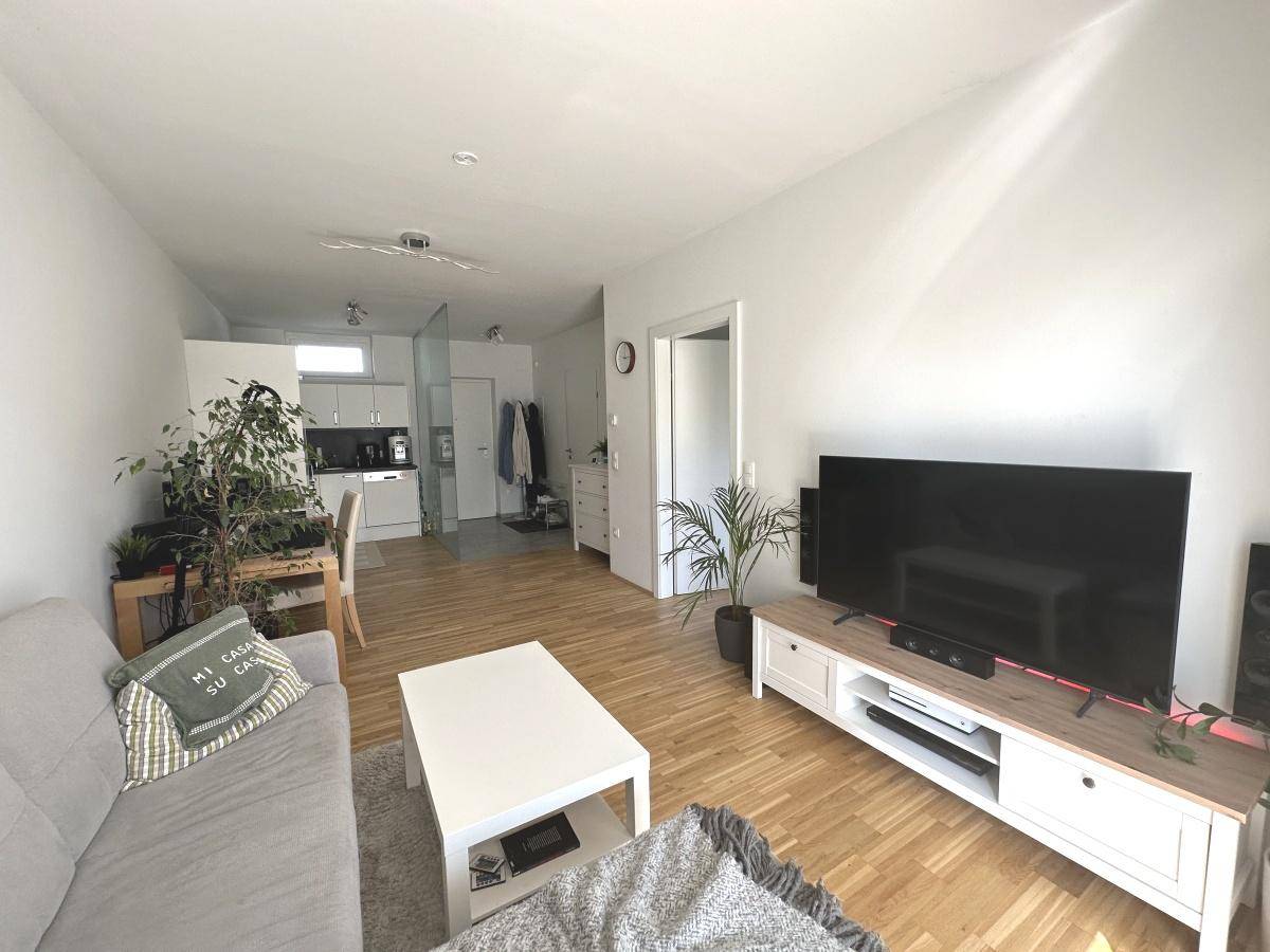 Heller Wohnbereich mit Holzboden, Sofa, Fernseher und offener Verbindung zur Küche.