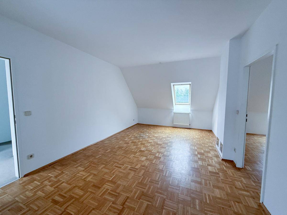 Großzügiger Wohnbereich mit warmem Parkettboden, weißen Wänden und einem Dachfenster, das Licht spendet.