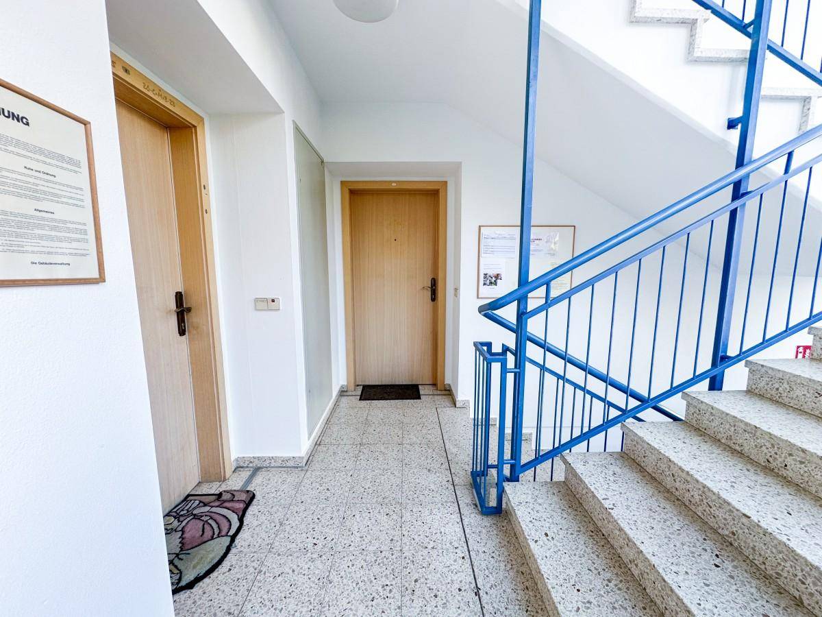 Helles Treppenhaus mit Terrazzoboden, blauen Geländern und Aufzugzugang.