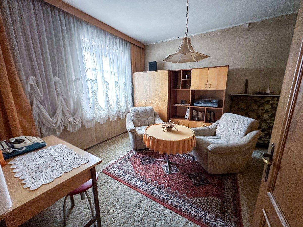 Zimmer mit Teppichboden, zwei Sesseln und einem kleinen Tisch am Fenster.