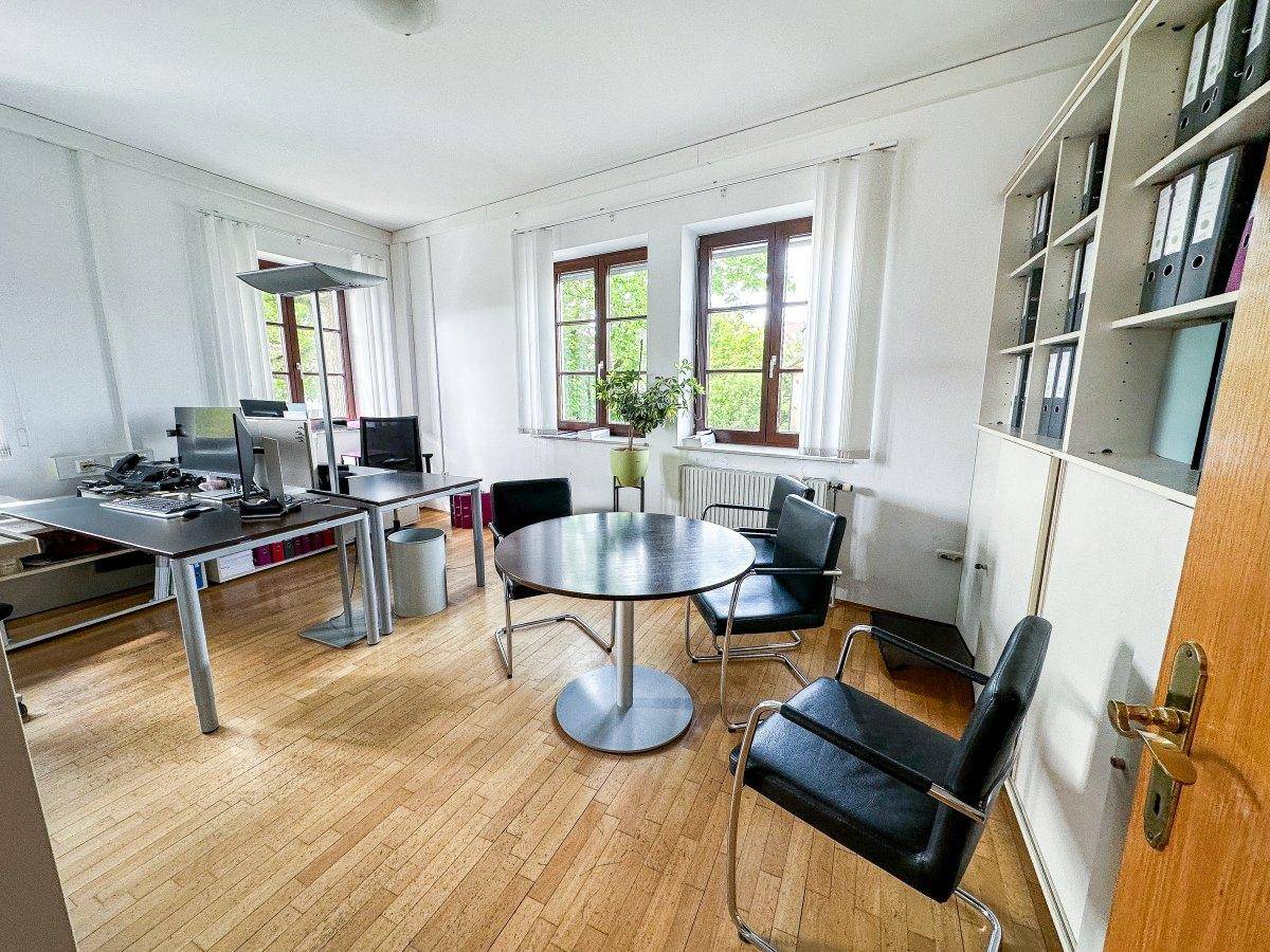 Geräumiges Büro mit zwei Fenstern, Holzfußboden und mehreren Arbeitsplätzen mit Computern.