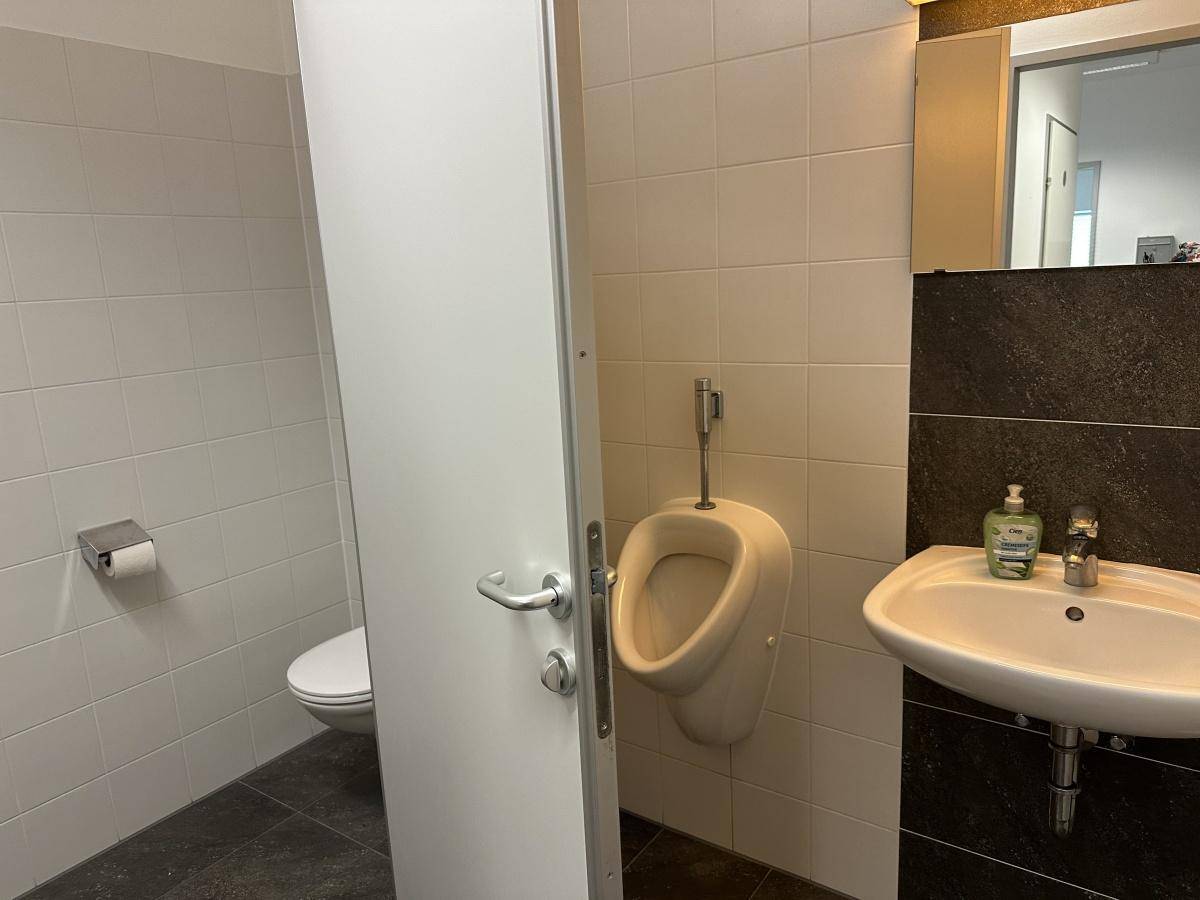 Herren-WC mit Toilette, Urinal und Waschbecken, ausgestattet mit weißen Wandfliesen und dunklem Boden.