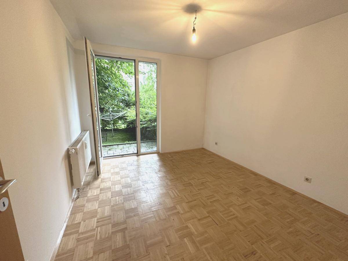 Geräumiges Zimmer mit Parkettboden und direktem Zugang zu einem grünen Außenbereich.