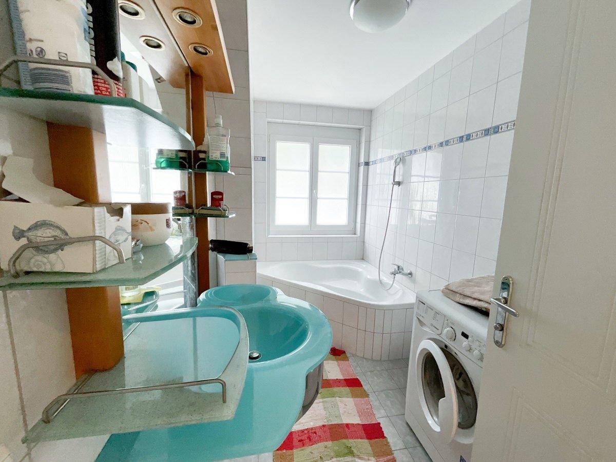Badezimmer mit Eckbadewanne, Waschmaschine und Fenster, hell gefliest.