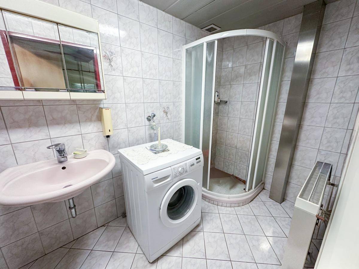 Badezimmer mit Duschkabine, Waschmaschine und hellen Fliesen an Wänden und Boden.
