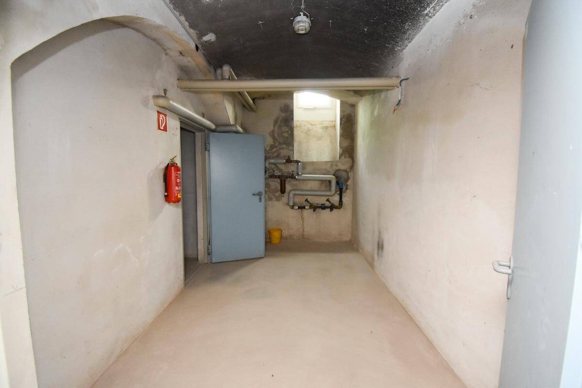 Kellerraum mit sichtbaren Rohren, Betonboden und einem Feuerlöscher an der Wand.