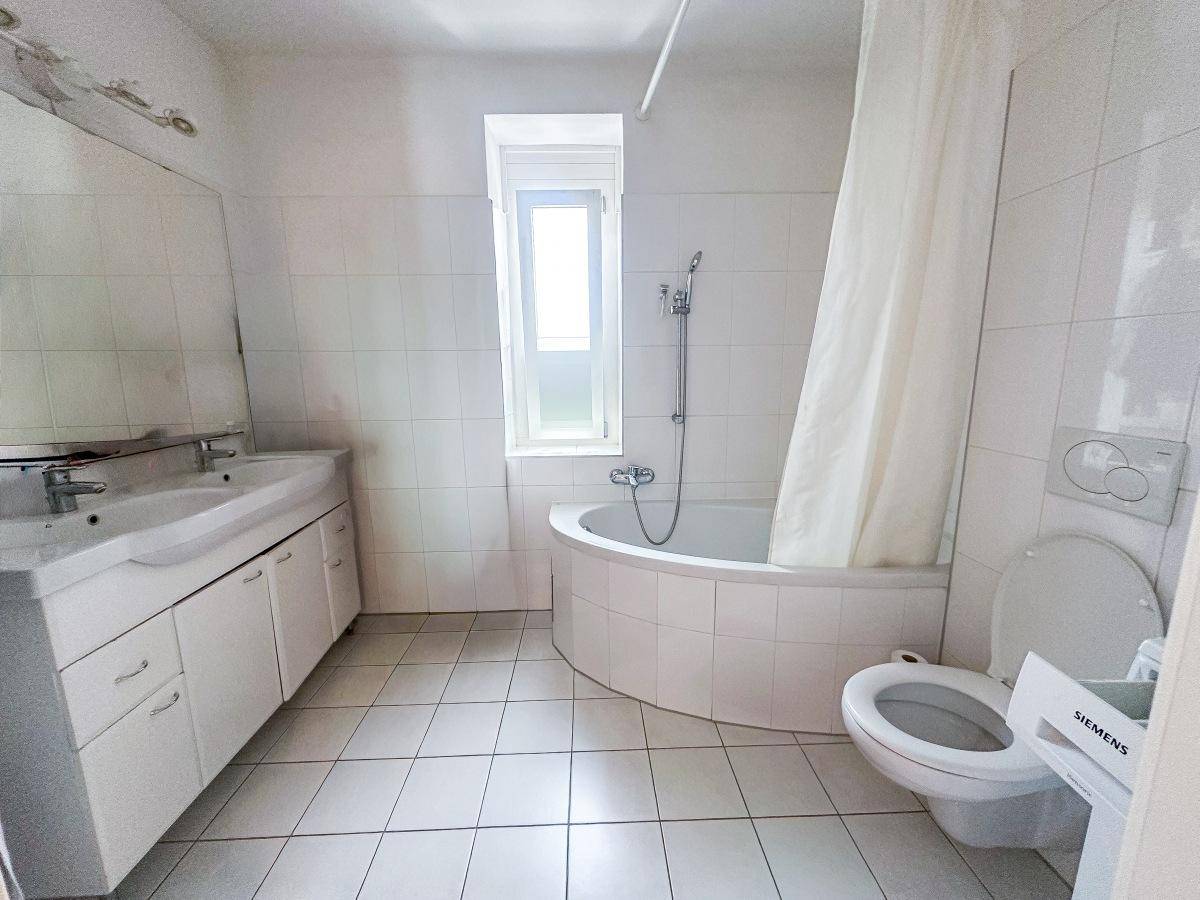 Badezimmer mit Doppelwaschbecken, Badewanne und Fenster, komplett weiß gefliest.