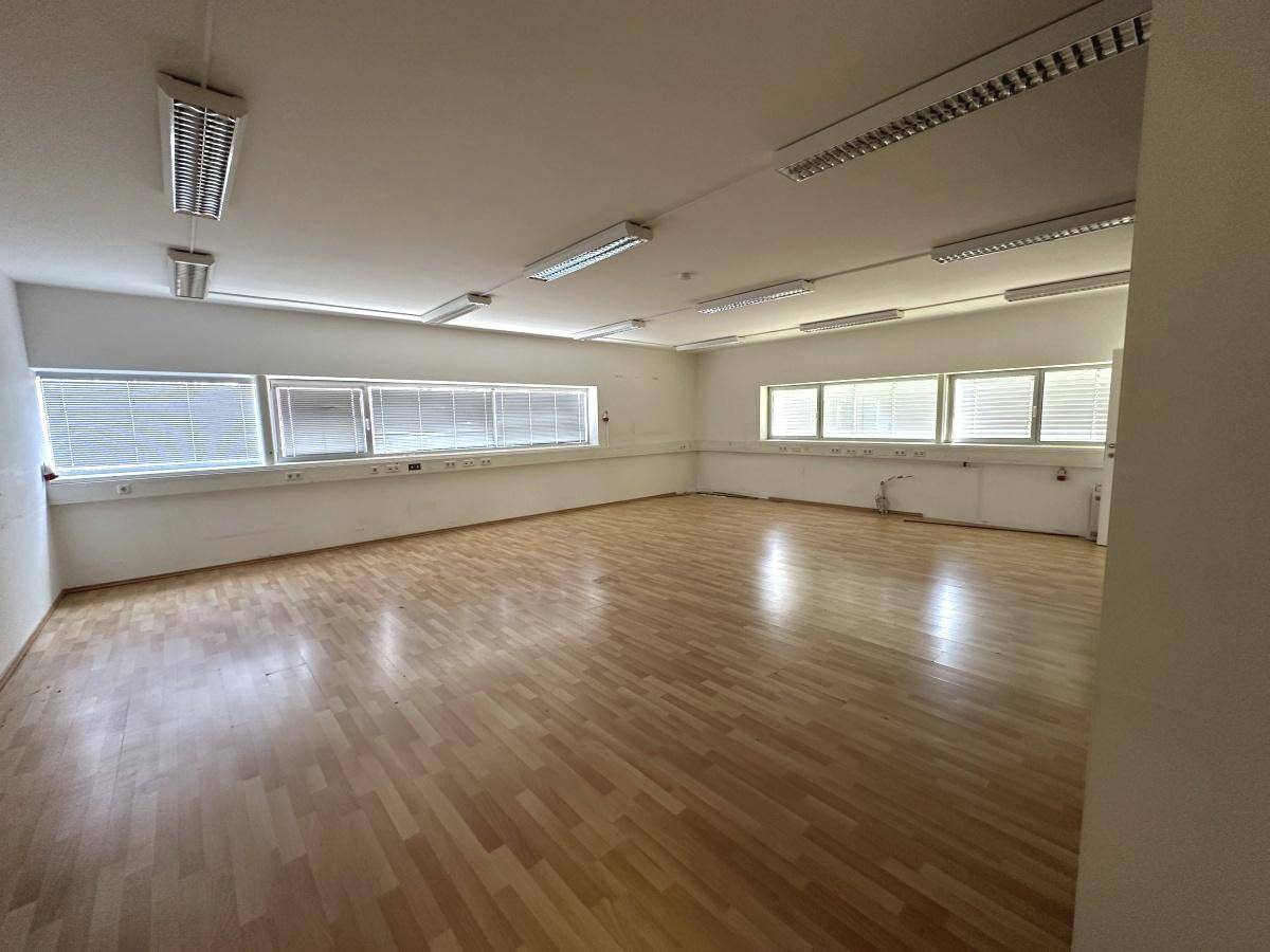Geräumiger, leerer Büroraum mit Holzparkettboden, vielen Fenstern und Deckenbeleuchtung.