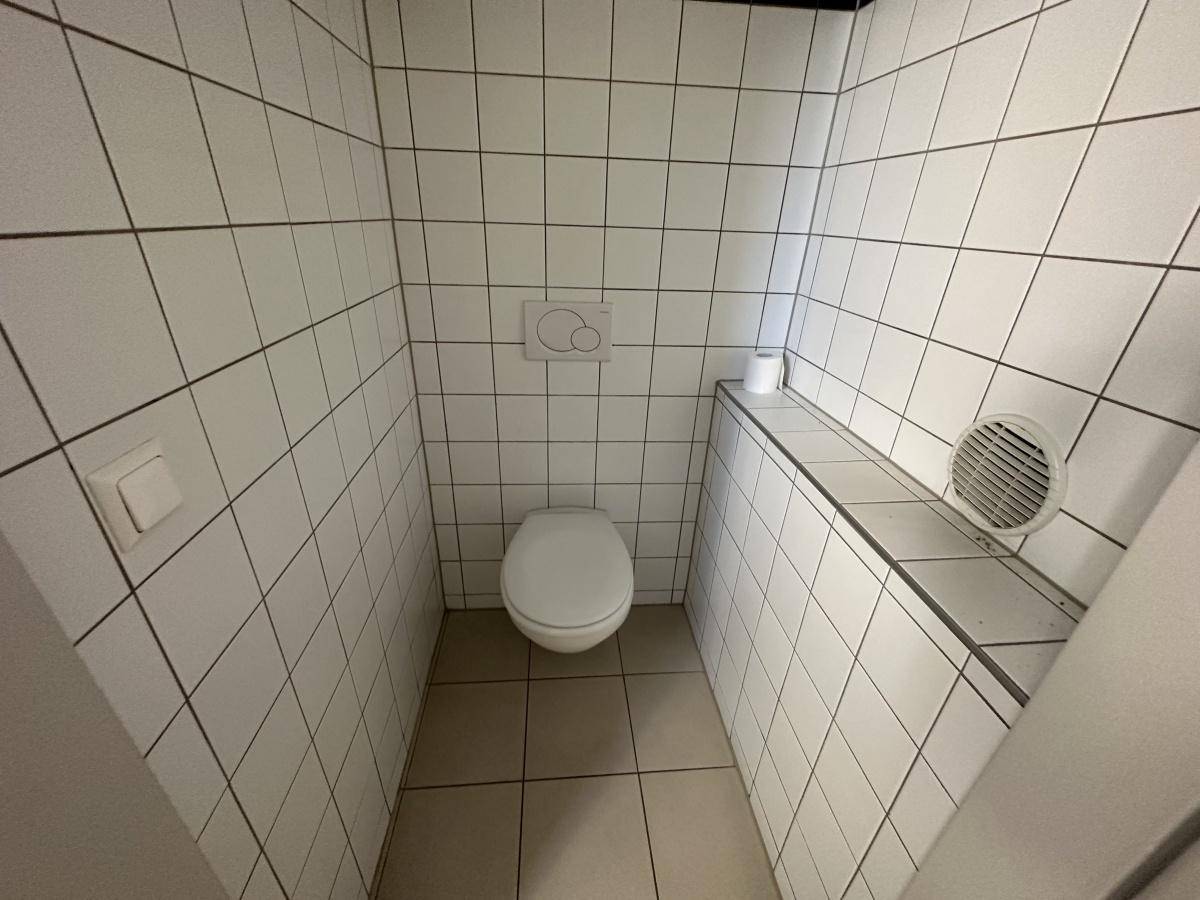 Toilette mit weißen Fliesen, WC und einem Abluftventilator an der Wand.
