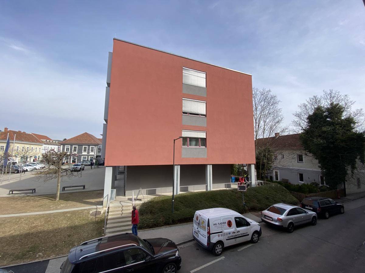 Moderne rote Gebäudefassade mit Fenstern und Jalousien, umgeben von einer Straße und Parkplätzen.