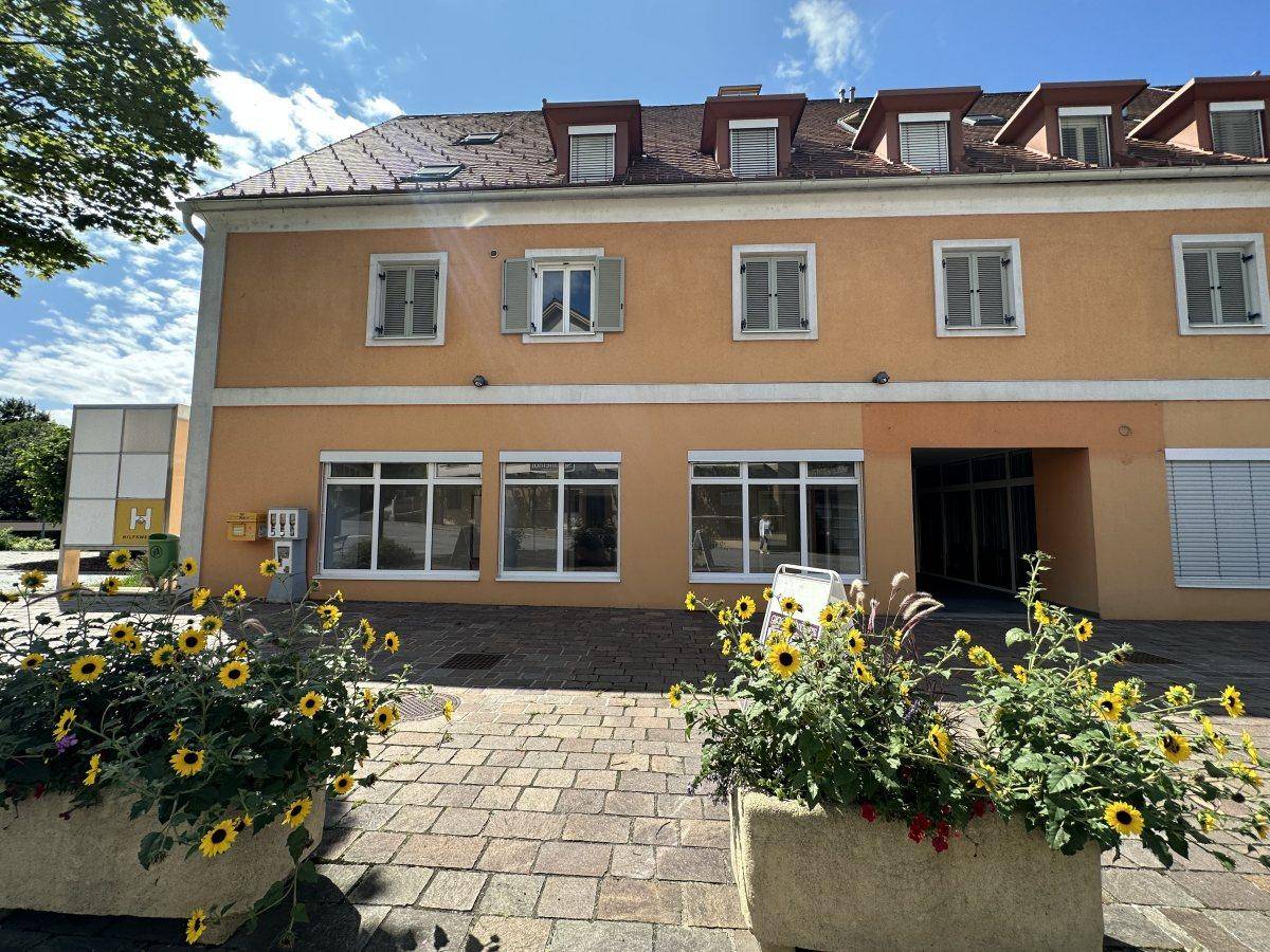 Gebäudeansicht mit orangefarbener Fassade, Fenstern mit Fensterläden und Blumenkübeln im Vordergrund.