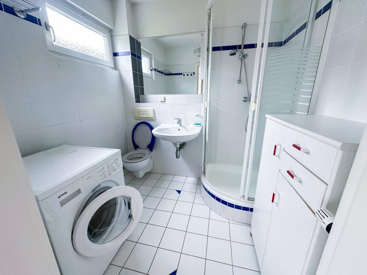 Funktionales Badezimmer mit Dusche, Waschbecken, WC und Waschmaschine in hellen Fliesen.