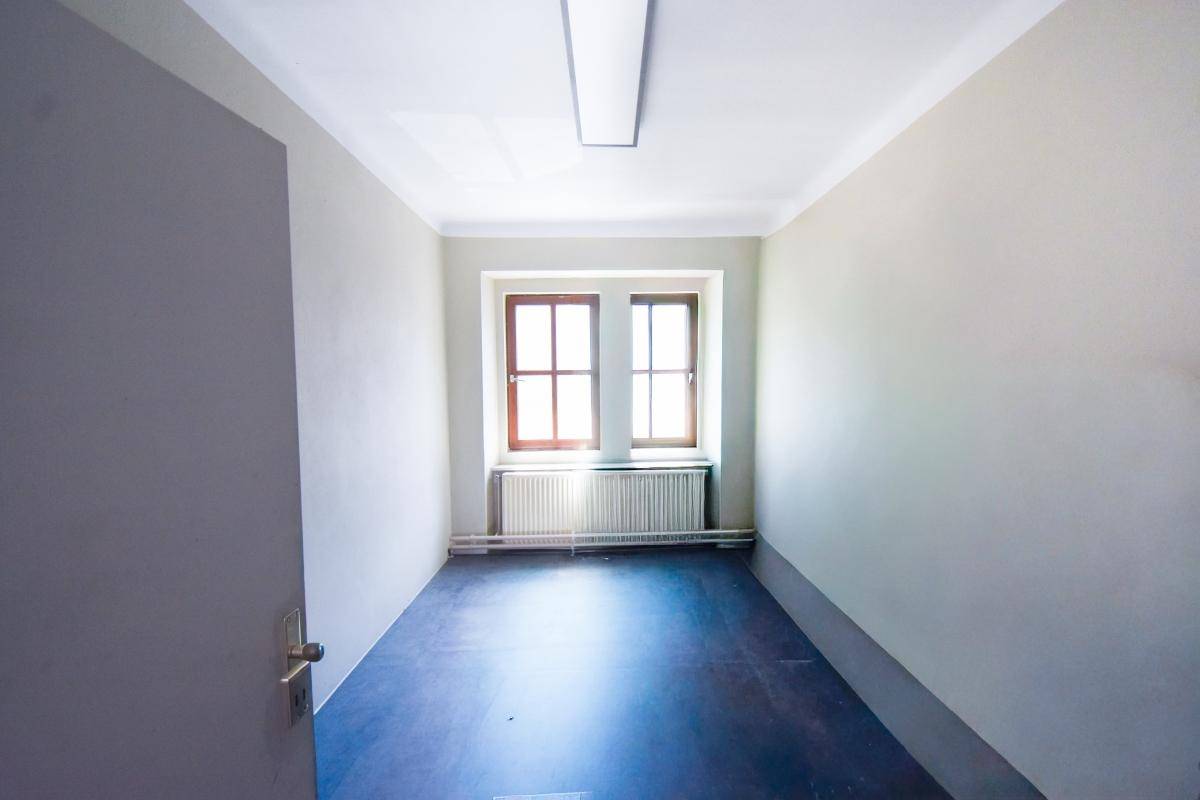 Schlichter, leerer Raum mit dunklem Bodenbelag, hellen Wänden und zwei Fenstern mit Heizkörpern.