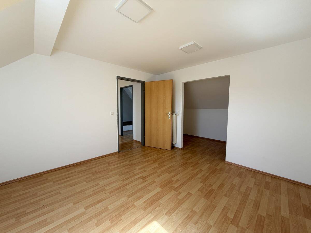 Leeres Zimmer im Dachgeschoss mit hellem Laminatboden und Zugang zu weiteren Räumen.